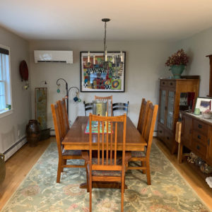 A dining room with a table and chairs center and the indoor ductless mini split head for a cold climate air source heat pump on the wall next to artwork.