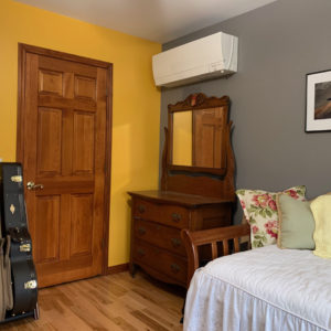 image of a bedroom with a bed, door, and antique washstand, with a Mitsubishi ductless mini split heat pump on the wall above the washstand.