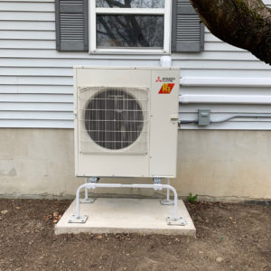 Shows a square tan metal rectangle on a stand with a fan and grill over the fan. It is an example of a Mitsubishi HyperHeat cold climate air source heat pump's outside heat exchanger.