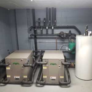 Shows two metal boxes, the size of large moving boxes, and a tank connected with piping. These are Geostar Ashton heat pumps, exchanging heat from the ground and increasing it to the temperature needed to heat the space.