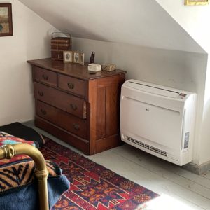 A cold climate air source heat pump low wall unit mounted under an eave next to a dresser in a bedroom