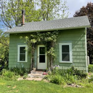 A small green house in upstate NY with gardens and an old wood stove pipe coming out of the roof that is now heated and cooled with cold climate air source heat pumps.