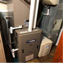 Shows a high efficiency gas furnace with the cold climate air source heat pump indoor heat exchanger in the metal box above it. Ductwork to the rest of the house can be seen beside it.