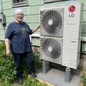 An image of an older homeowner smiling with the outdoor units for her cold climate air source heat pump